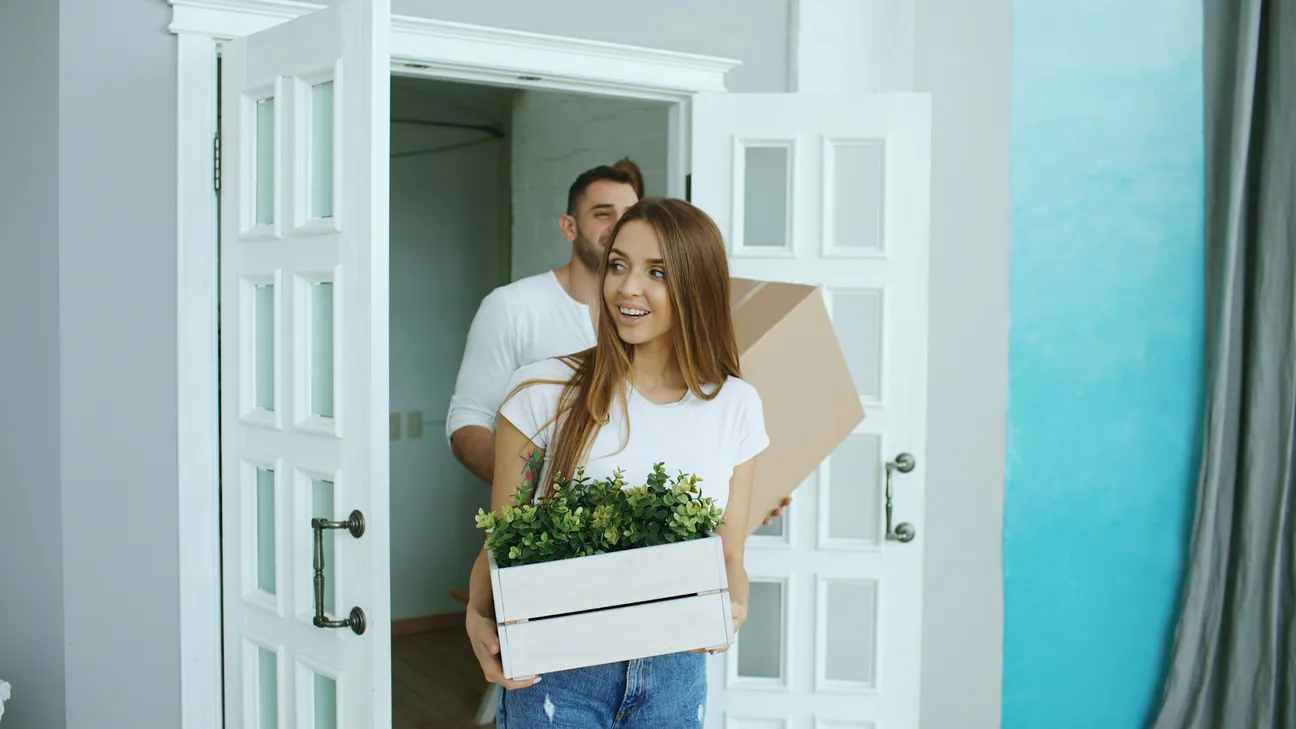 A couple carrying a box and a plant crate through the front door of a home